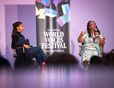 Two women sit on stage having a discussion in front of a banner that reads “PEN America World Voices Festival.” One woman holds a microphone while the other listens attentively. The background is purple.