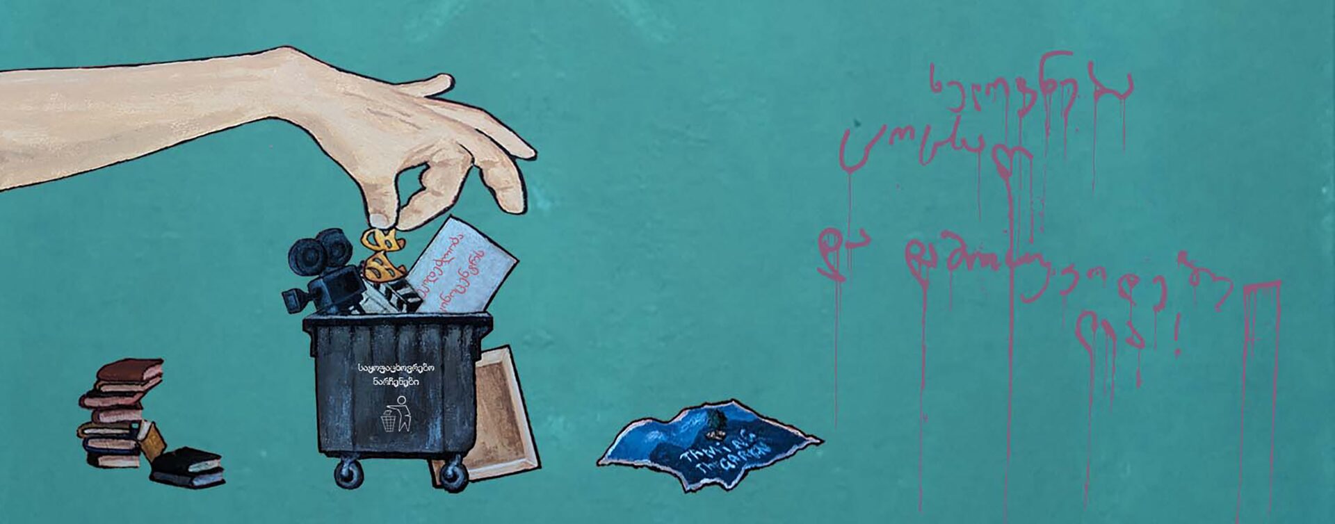 A painting shows a hand dropping a camera, books, and a paper labeled freedom of speech into a trash bin—an image reflecting the taming culture in Georgia. More books lie by the bin, with red Georgian writing on the green wall beside the scene.