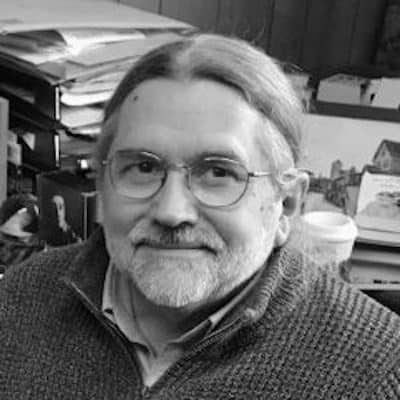 A middle-aged man with long hair and glasses, possibly a Pen America Literary Awards nominee, smiles at the camera. He wears a sweater and sits before a cluttered desk filled with books and papers in this black-and-white photo.