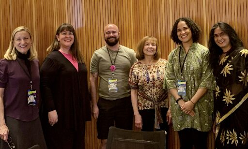 Six people stand side by side indoors, smiling at the camera. They wear a mix of casual and semi-formal outfits and conference badges, with a wooden paneled wall in the background.