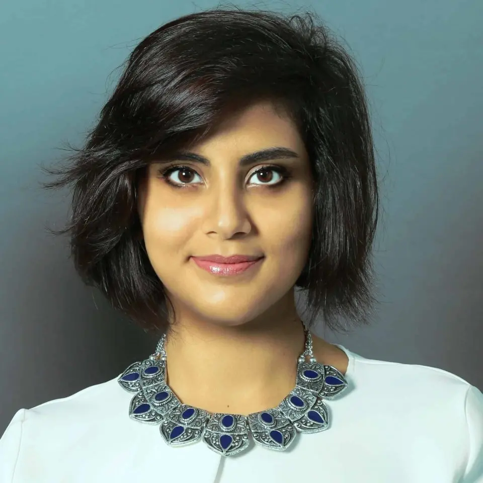 A woman with short, dark hair and brown eyes smiles slightly. She is wearing a white top and a large, ornate silver necklace with blue accents, standing before a plain grey background—her confidence echoing the spirit of the Freedom to Write Index 2019.