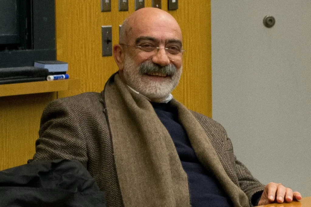 A smiling older man with glasses, a bald head, and a gray beard, wearing a brown blazer, scarf, and dark sweater, sits indoors near a wooden cabinet—a thoughtful presence reminiscent of the Freedom to Write Index 2019.