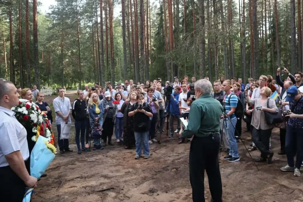 A large group of people stands in a wooded area, listening to a man address them. Some hold flowers and cameras, suggesting a commemorative event linked to the Freedom to Write Index 2019. Tall pine trees surround the scene.