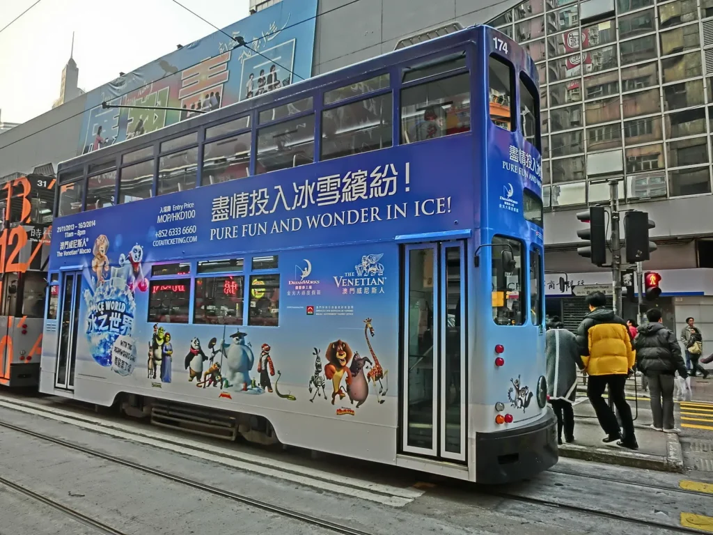 A double-decker tram in Hong Kong, featured in the China Hollywood Report, is decorated with animated penguin characters and blue graphics for Pure Fun and Wonder in Ice. People walk nearby on the street, with buildings in the background.