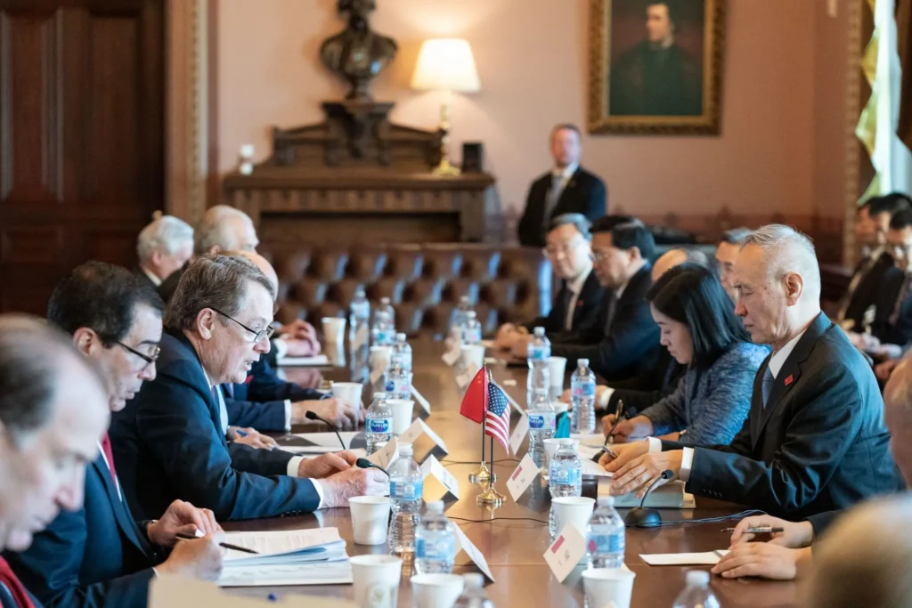 A group of officials from two delegations, including representatives from the China Hollywood Report, sit across from each other at a long conference table with documents and flags, engaged in a formal meeting in an ornate room.
