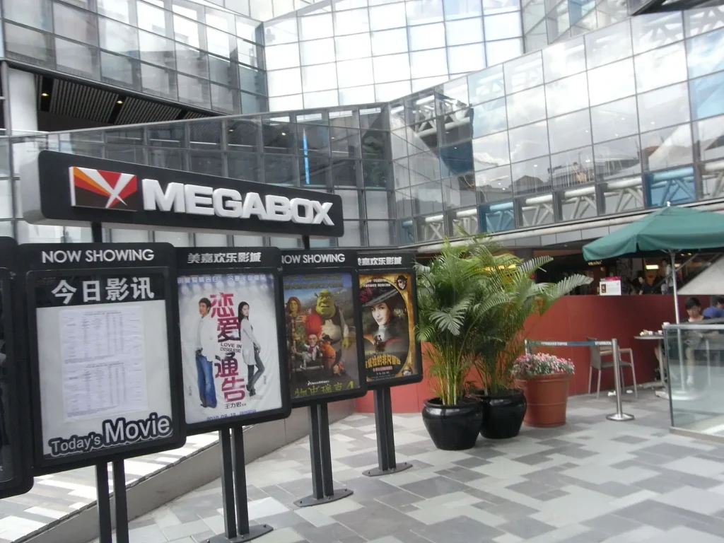 Outdoor view of a MEGABOX cinema, featured in the China Hollywood Report, with a movie listing board showing film posters, surrounded by glass buildings, potted plants, and people near a café area.