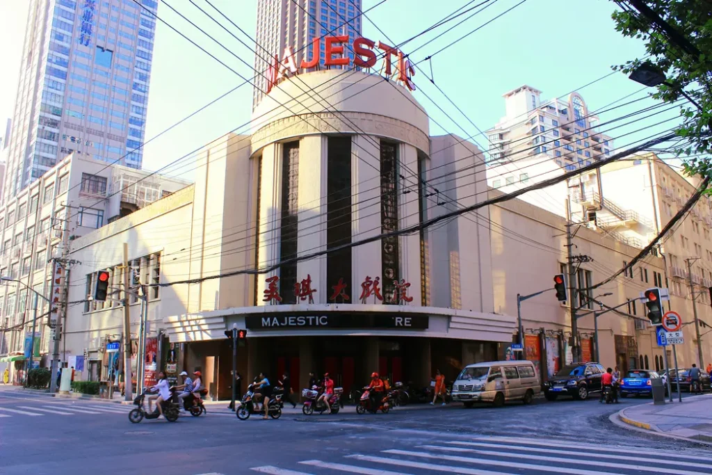 A street view of the Majestic Theatre in Shanghai, captured like a scene from a China Hollywood report, with people on scooters and cars waiting at the intersection. The tall art deco building features vertical windows and red Chinese characters above its entrance.