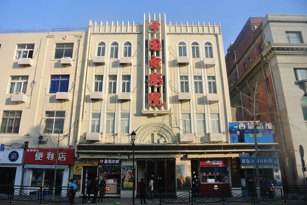 A beige Art Deco-style building with balconies and a large vertical sign in Chinese characters, reading China Cinema, stands between shops and businesses on a sunny street—a scene worthy of a China Hollywood report.