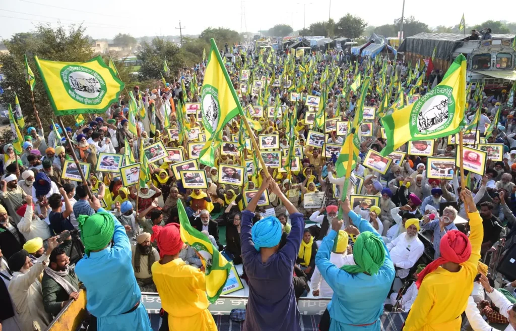 A large crowd gathers outdoors, many waving yellow and green flags and holding up framed photos. Several people in colorful outfits and turbans stand on a platform, rallying support for digital sovereignty among the enthusiastic supporters below.