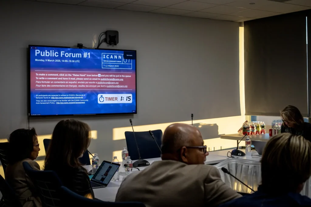 A group of people sit at a conference table with laptops during a meeting focused on digital sovereignty. A screen displays information for Public Forum #1 by ICANN, sunlight streams through a window, and drinks are arranged on a side table.