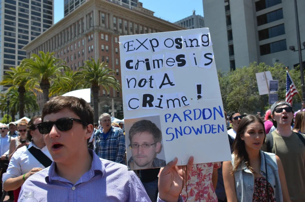 People march in a city protest holding a sign that reads, Exposing crimes is not a crime! Pardon Snowden, with a photo of Edward Snowden. The demonstration highlights calls for digital sovereignty amid palm trees and tall buildings in the background.