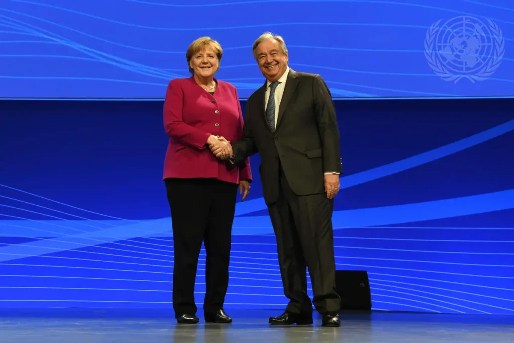A woman in a red blazer and a man in a dark suit stand on stage in front of a blue backdrop with UN logos, smiling and shaking hands at an event highlighting digital sovereignty.