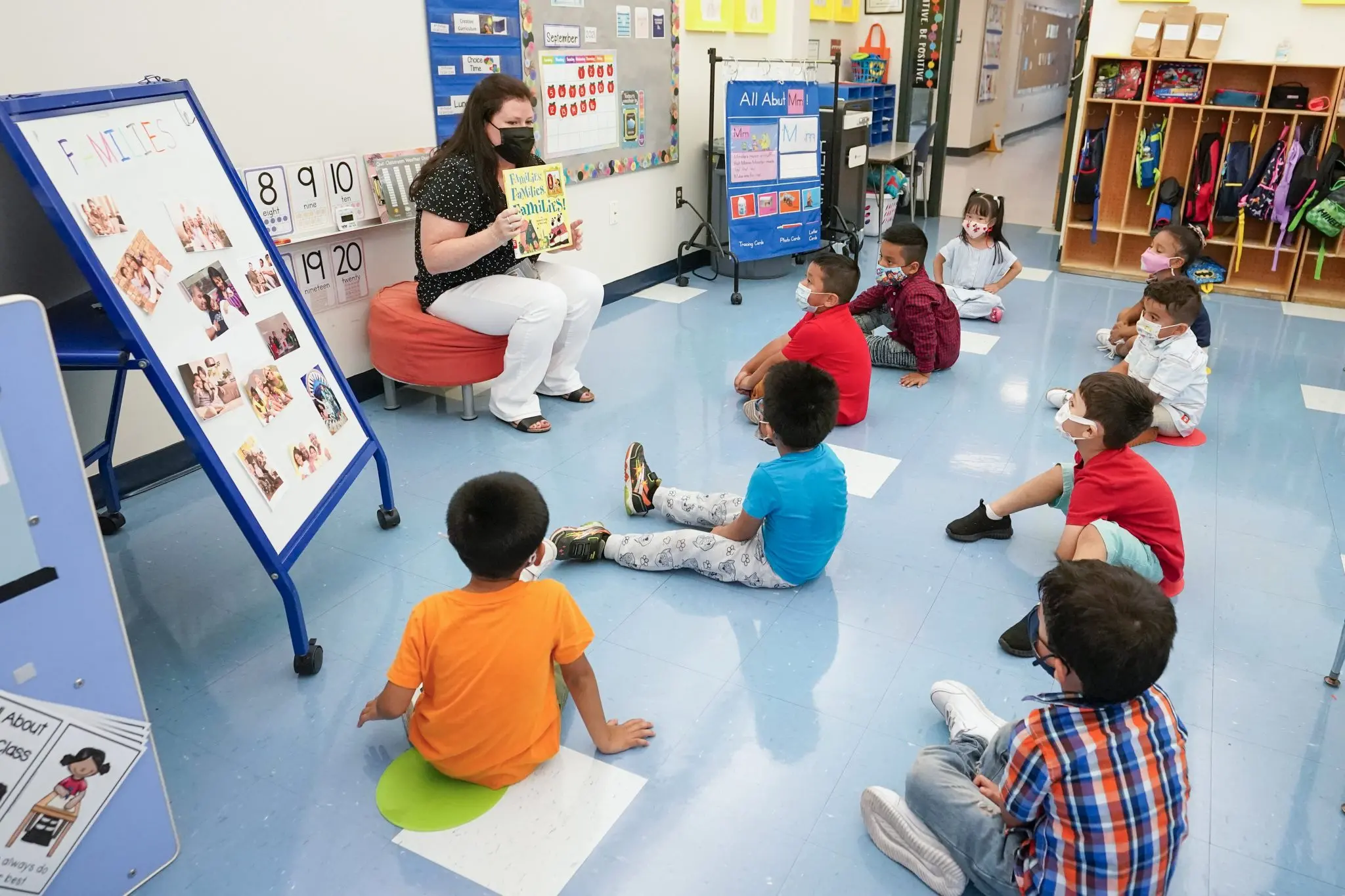 A teacher wearing a mask reads a book to young children, also masked, seated on the floor in a colorful classroom with educational posters and cubbies visible, despite recent concerns over Educational Gag Orders.