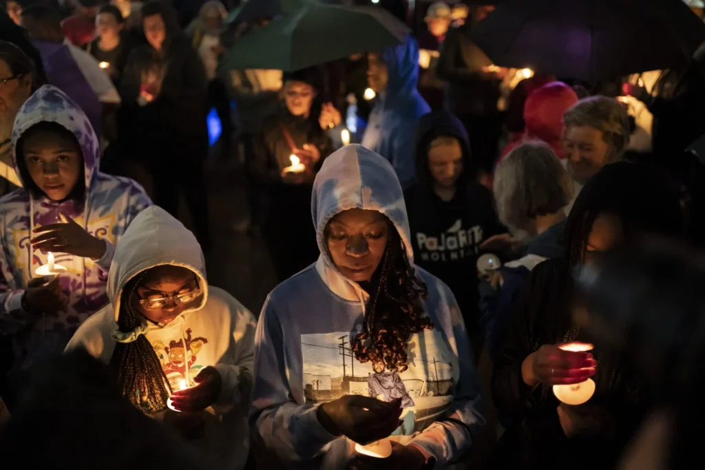 A diverse group of people stand close together at night, holding lit candles and wearing hoodies. Some look down in reflection about educational gag orders, while others look ahead. Several hold umbrellas, suggesting it may be raining.