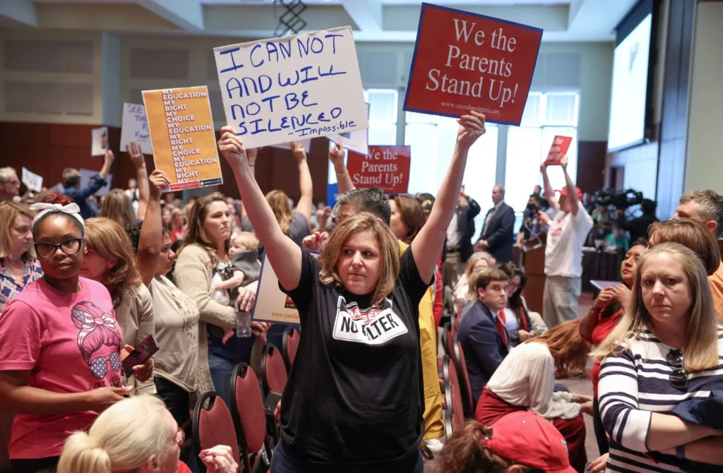 A woman in a crowd holds a sign reading I CAN NOT AND WILL NOT BE SILENCED, while others protest Educational Gag Orders with signs saying We the Parents Stand Up! at a public meeting, people seated and standing around her.