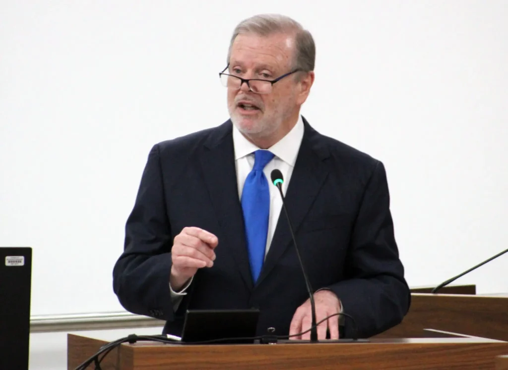 A man in a dark suit, blue tie, and glasses speaks at a wooden podium with a microphone, addressing the topic of Educational Gag Orders. He gestures with his right hand and looks focused. The background is plain and white.