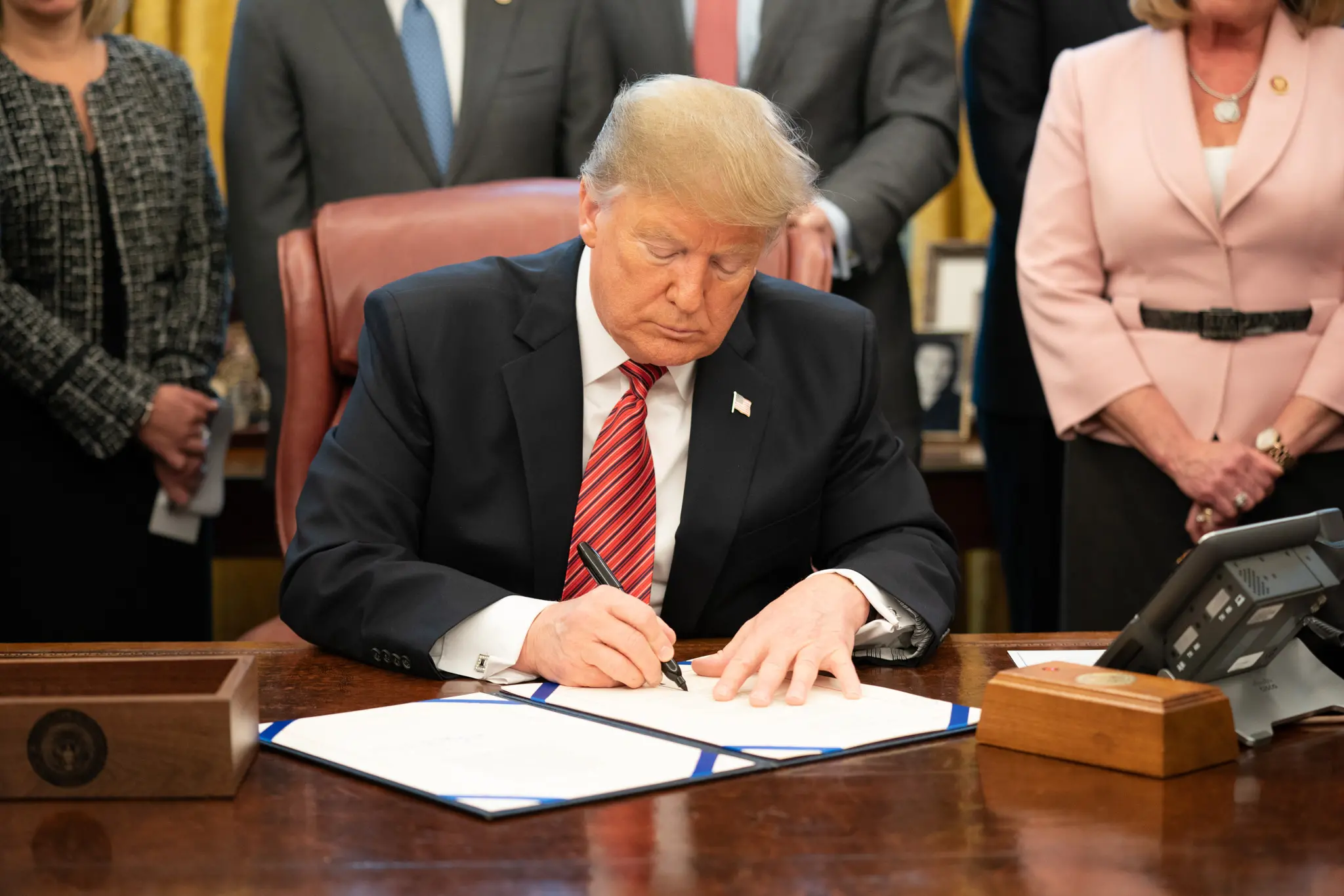 A man in a suit and red striped tie sits at a desk, signing an Educational Gag Order as several people in business attire stand behind him. The setting appears to be an official office.