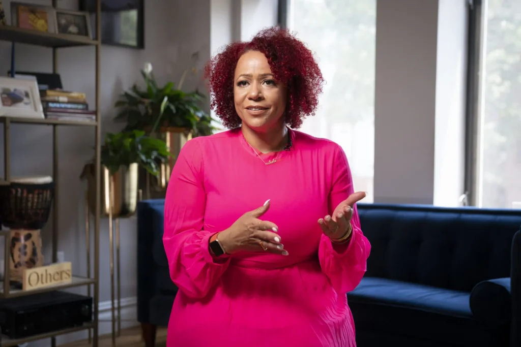 A woman with curly red hair wearing a bright pink dress gestures with her hands while seated on a blue sofa in a well-lit, modern living room, discussing the impact of Educational Gag Orders amid plants and books in the background.