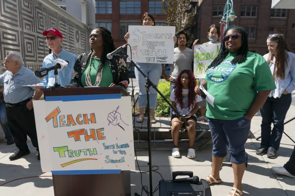 A diverse group gathers outdoors for a rally against Educational Gag Orders. A woman speaks at a podium with a sign reading "Teach the Truth," while others hold signs, including one that says, "Students deserve the truth.