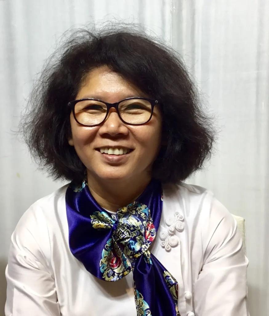 A woman with shoulder-length dark hair and glasses smiles at the camera. She is wearing a white blouse with decorative buttons and a blue, floral-patterned scarf tied around her neck, reflecting hope after the Myanmar 2021 coup. The background is a white curtain.
