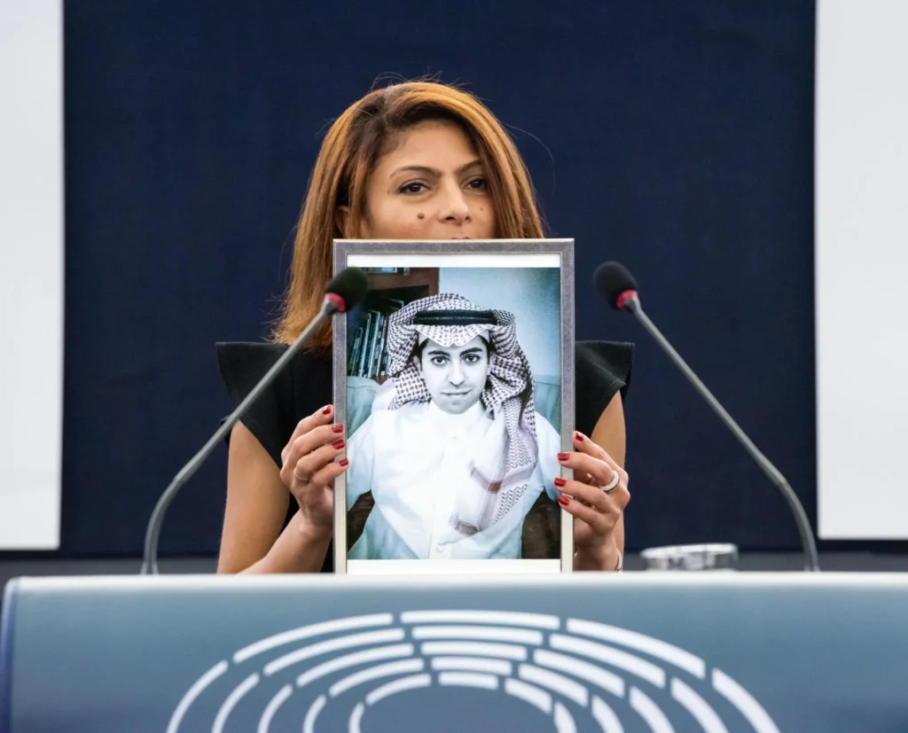 A woman stands at a podium with microphones, holding up a framed black-and-white photo of a man in traditional Middle Eastern attire, possibly highlighting findings from the Freedom to Write Index 2021 at an official event.