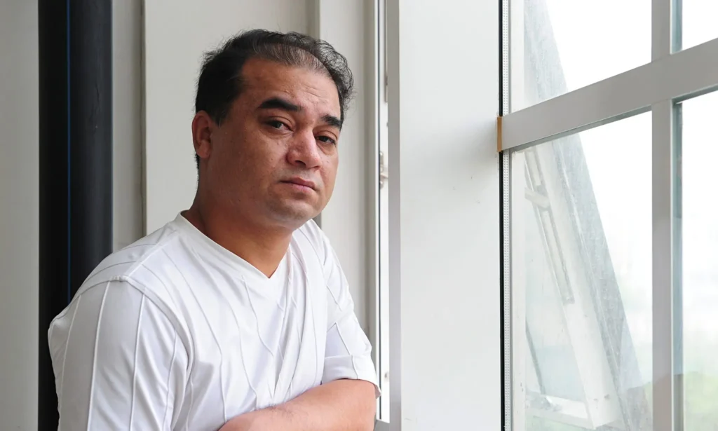 A man with short dark hair, wearing a white shirt, stands indoors and looks thoughtfully out of a large window with crossed arms. Natural light brightens the room, evoking the contemplative spirit of the Freedom to Write Index 2021.
