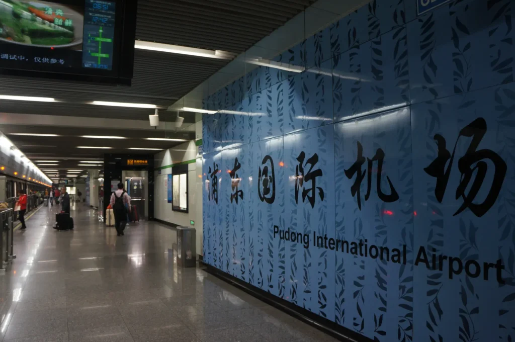 A hallway in a subway or train station with a large blue wall sign reading Pudong International Airport in English and Chinese. Travelers with luggage walk along the corridor under bright lighting, reminiscent of scenes from the Freedom to Write Index 2021.