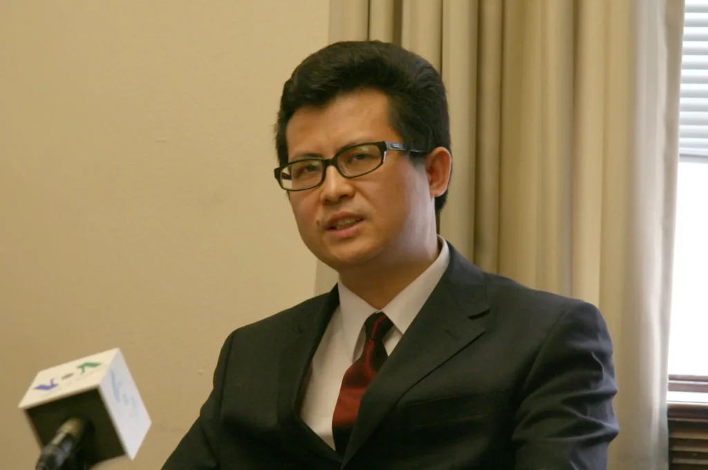 A man in a dark suit, red tie, and glasses sits at a table speaking into a microphone. The scene, set against beige curtains and a window, evokes the spirit of discussions highlighted in the Freedom to Write Index 2021.