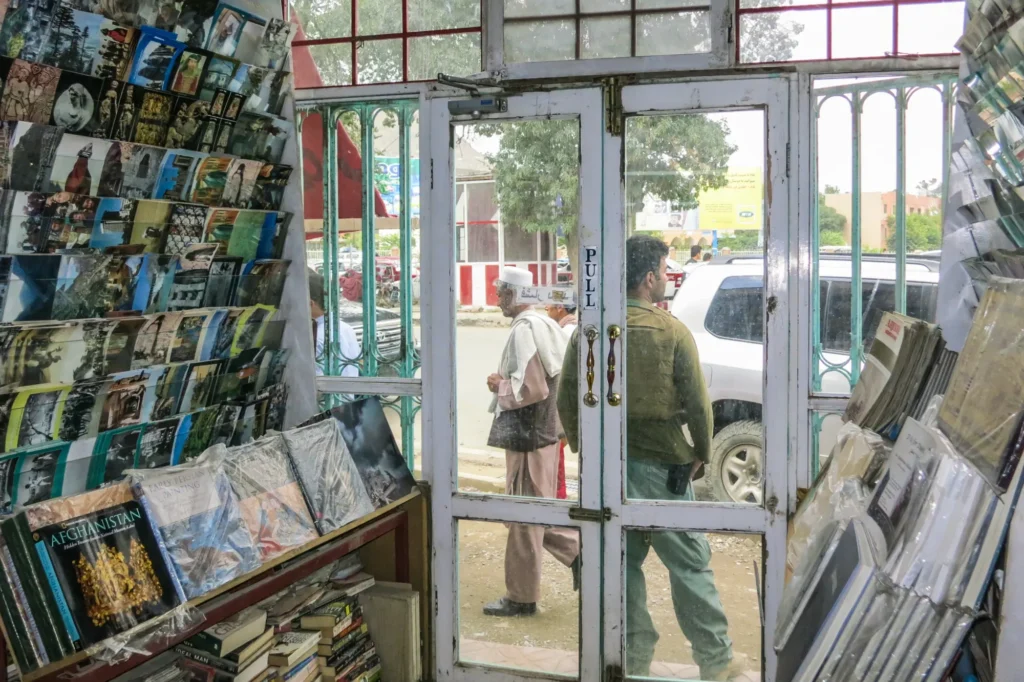 View from inside a bookstore with shelves of books and magazines. Through the glass door, two people walk outside on a sidewalk—cars and trees beyond—reflecting the spirit of expression celebrated in the Freedom to Write Index 2021.