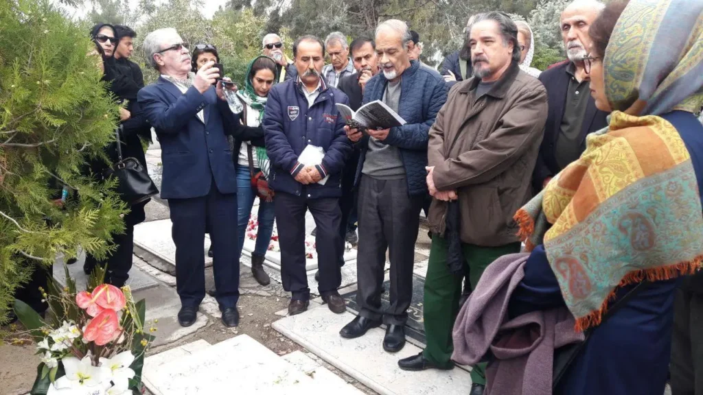 A group of people stands together at a cemetery, some holding flowers and books, possibly honoring a writer listed in the Freedom to Write Index 2021. One man reads aloud while others listen solemnly among trees and gravestones.
