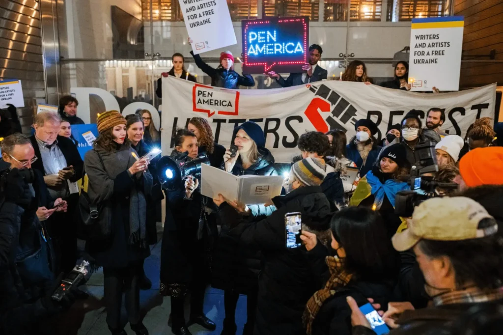 A crowd gathers at a PEN America event, holding signs that read Writers Resist and Writers and Artists for a Free Ukraine. As the Freedom to Write Index 2021 is highlighted, speakers address the group while people take photos and listen attentively.