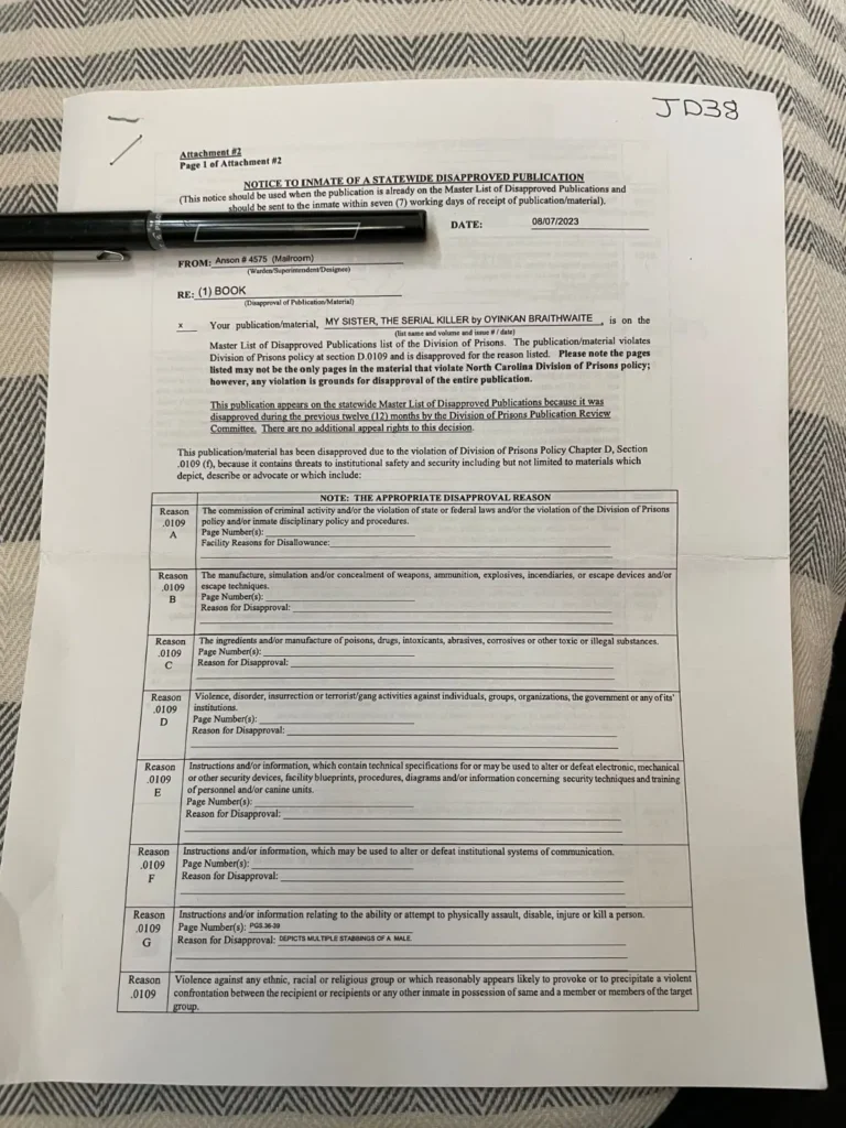 A printed notice titled Notice to Inmate of a Statewide Disapproved Publication lies on a striped fabric surface with a black pen across the top—an unexpected barrier to reading between the bars, as it concerns the book My Sister, the Serial Killer.