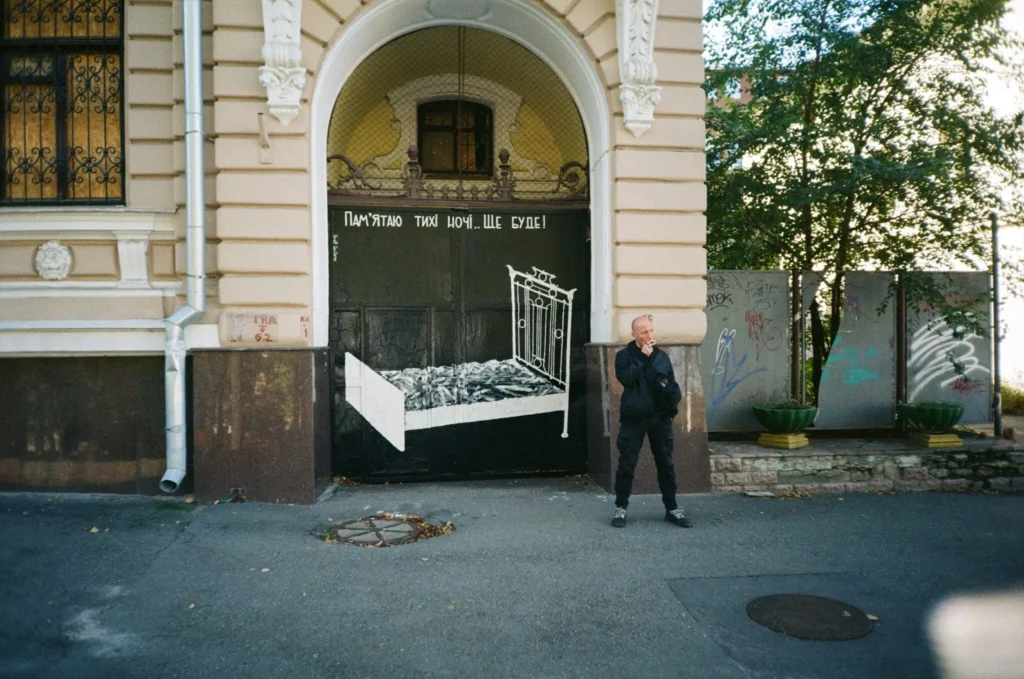 A man stands in front of a large arched doorway adorned with street art of a bed and Cyrillic writing, reflecting Ukrainian culture on the exterior of an ornate building with pale walls and ironwork. Trees and graffiti appear in the background.