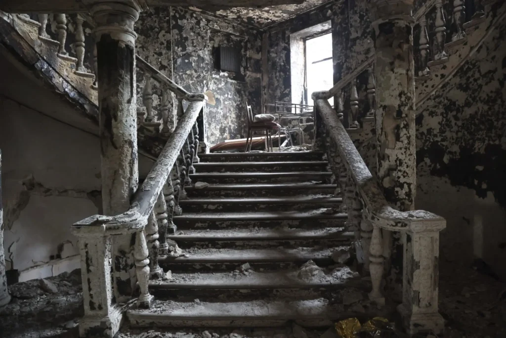 A grand staircase in a dilapidated, fire-damaged building, with peeling paint, debris on the steps, and broken railings. Two chairs and a table are visible at the landing near a window with light shining through.