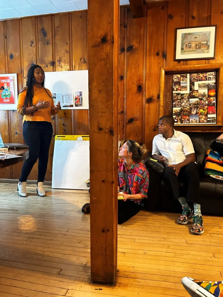 A woman stands and speaks near a whiteboard, discussing fighting disinformation locally, while two people listen attentively on a couch in a wood-paneled room. A large wooden pillar is in the center of the image.