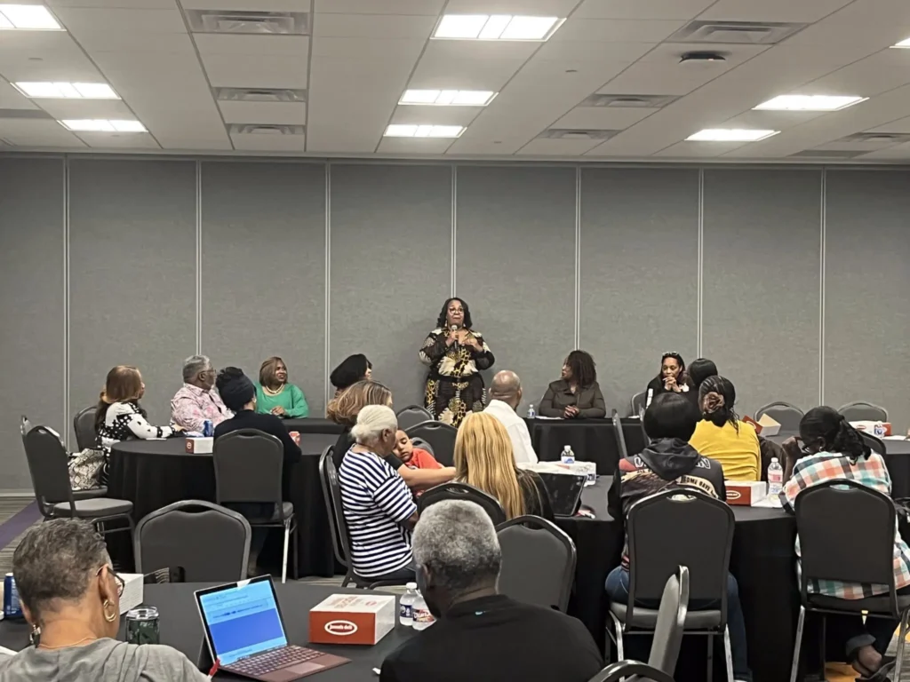 A woman stands speaking at the front of a conference room, addressing people seated at round tables about fighting disinformation locally. The attendees listen attentively, some with open laptops and notepads on the tables.