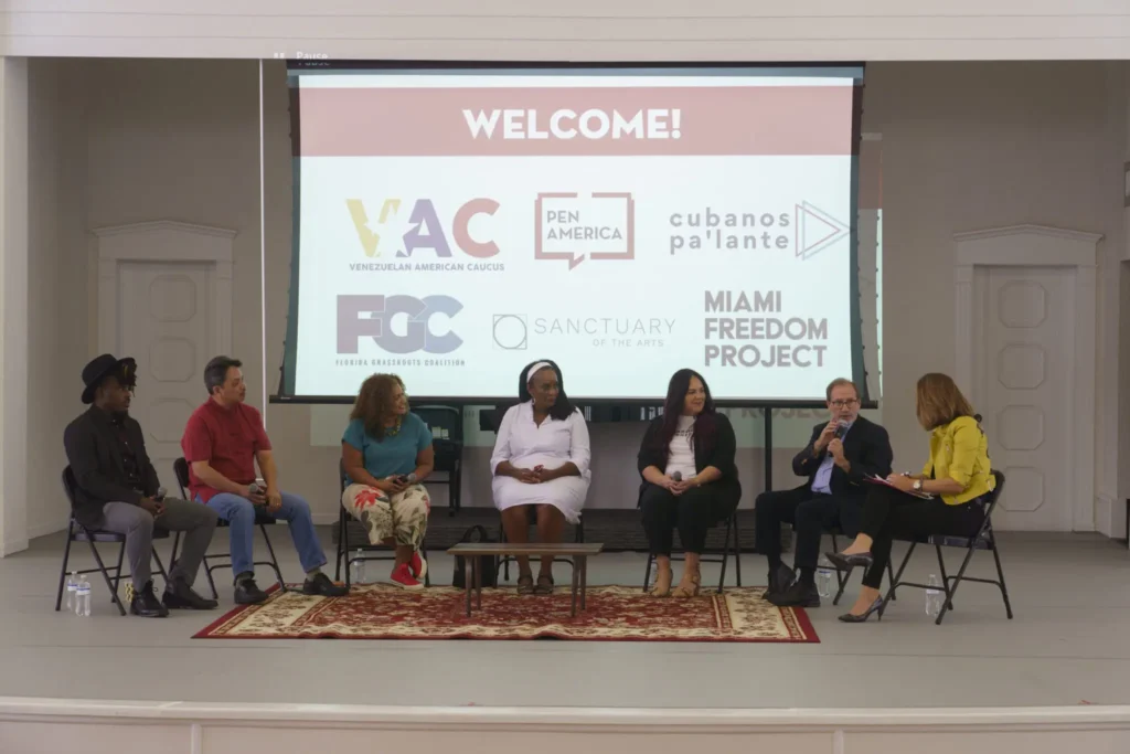 Six people sit on a stage in front of a screen displaying logos and the word “WELCOME!” They appear to be participating in a panel discussion about fighting disinformation locally, set in an event space with white walls and a decorative rug.