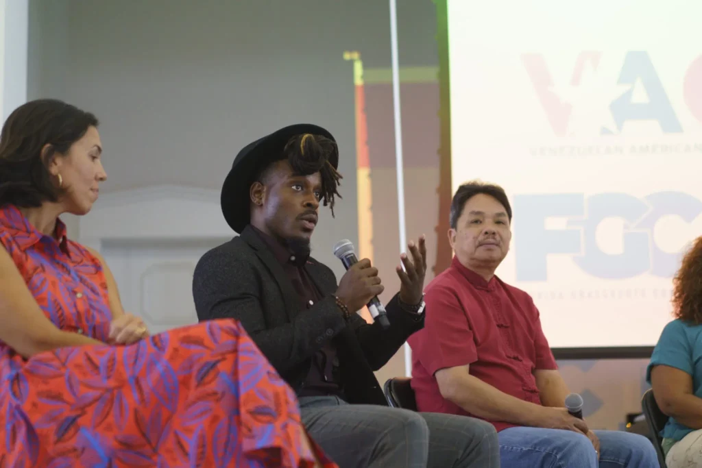 Four people sit on a panel; one person in a dark jacket and hat speaks into a microphone about fighting disinformation locally, while the others listen. A presentation screen with logos appears in the background.