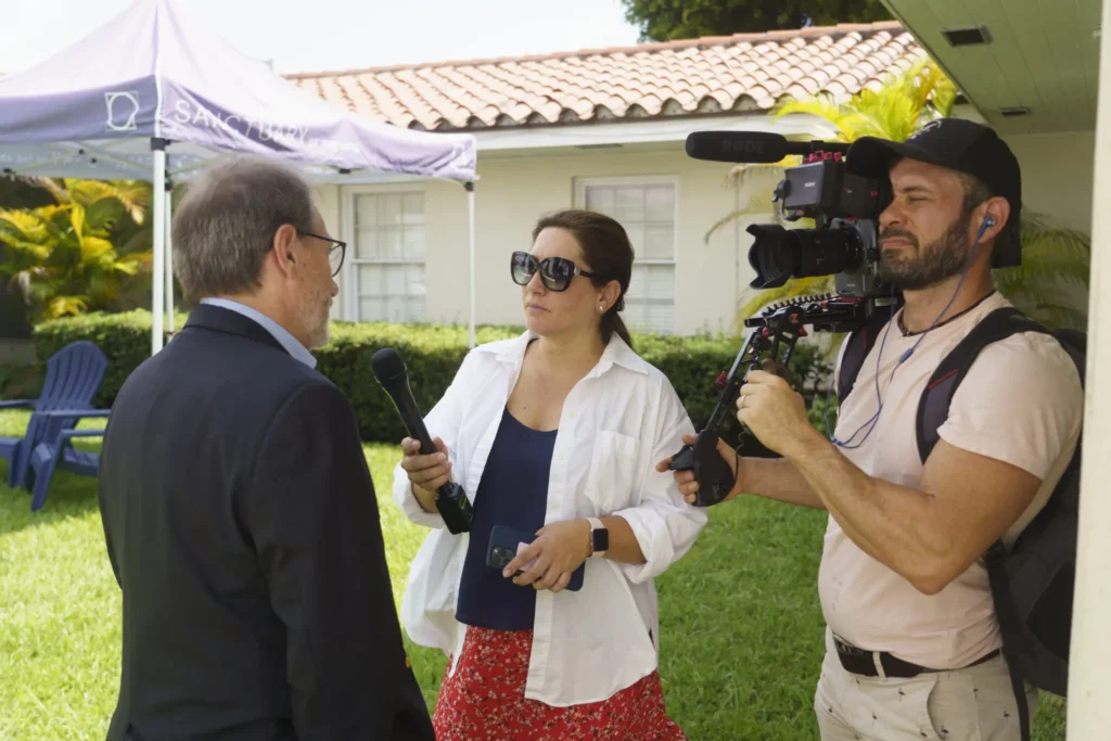 A woman holding a microphone interviews a man in a suit outdoors while a cameraman films them; they stand near a house and lawn on a sunny day, discussing ways of fighting disinformation locally.