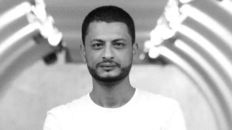 A man with short hair and a beard, wearing a plain white t-shirt, stands indoors in front of a blurred background featuring curved, illuminated lines, evoking the atmosphere of the Freedom to Write Index 2022.