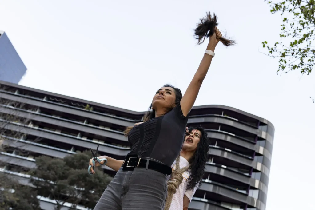 A woman raises a cut bundle of hair in one hand and scissors in the other, standing confidently in front of a modern building—a powerful symbol of change, echoing the spirit seen in the Freedom to Write Index 2022. Another woman smiles behind her.