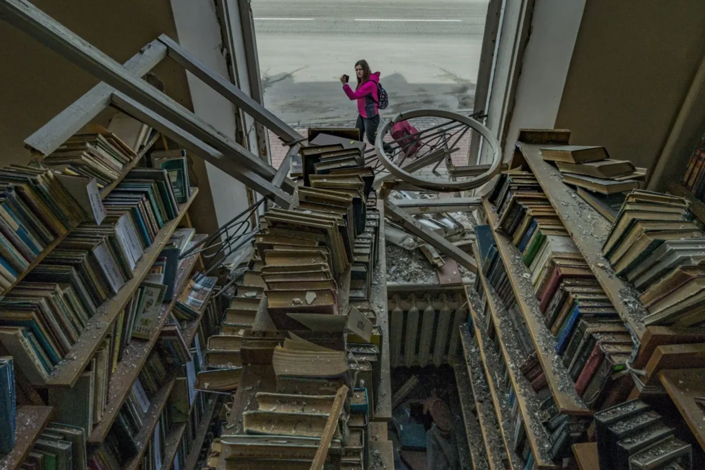 A woman in a pink jacket stands in the damaged remains of a library, surrounded by debris and leaning bookshelves—a scene reflecting the challenges highlighted in the Freedom to Write Index 2022.