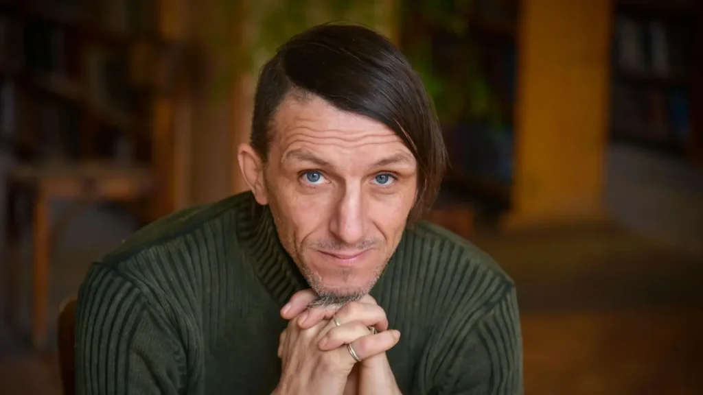 A man with blue eyes, wearing a green ribbed sweater, sits indoors with his hands clasped under his chin, looking directly at the camera. The softly blurred background with bookshelves hints at themes from the Freedom to Write Index 2022.