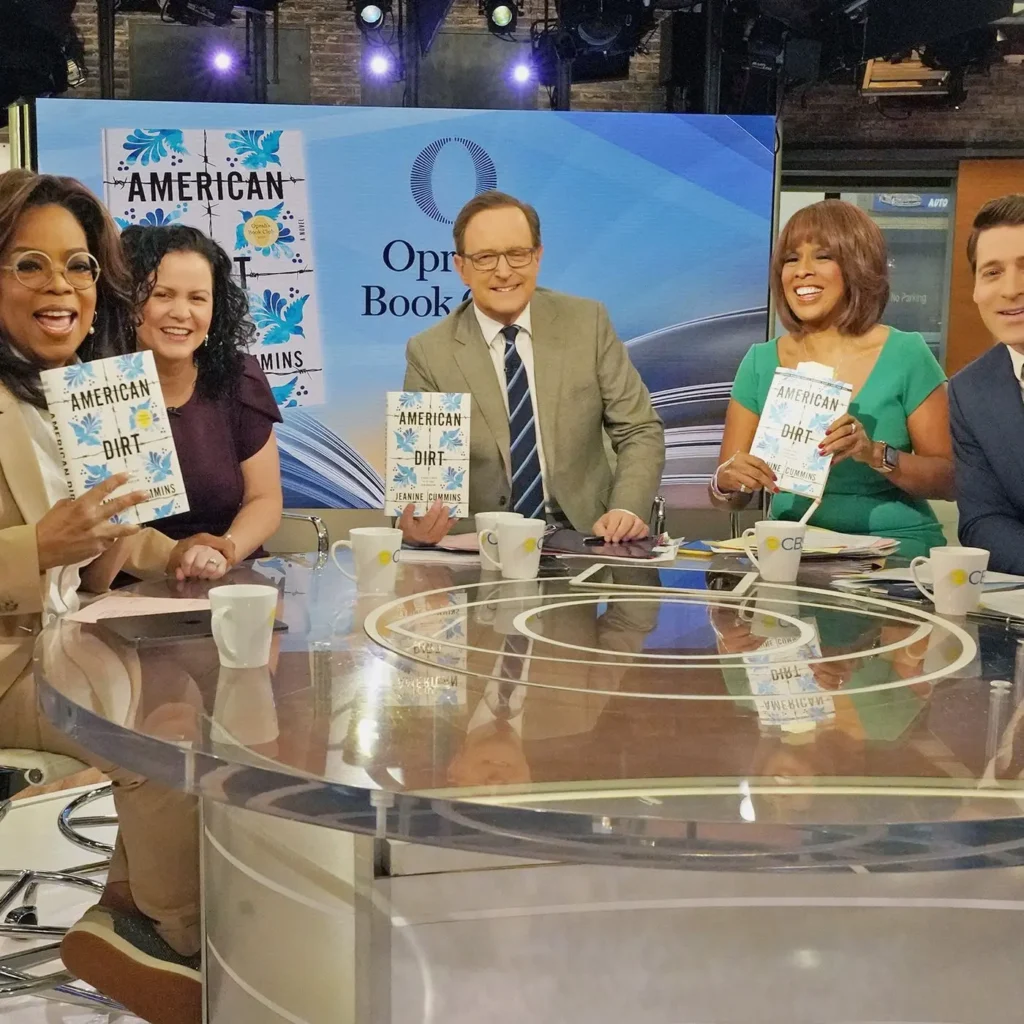 Four TV hosts sit at a news desk smiling and holding copies of the book American Dirt. The set displays the Oprah’s Book Club logo and book covers on screens, referencing the Booklash that sparked national discussion.