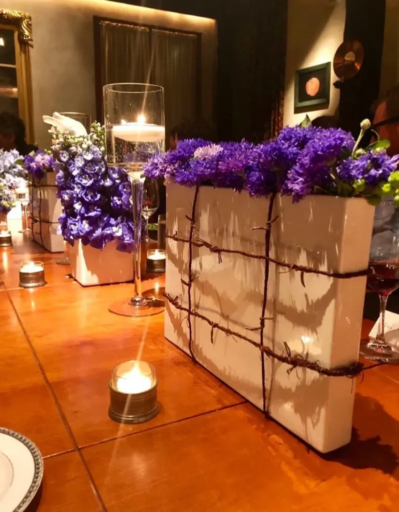 A warmly lit dinner table features rectangular white vases wrapped in twine and filled with vibrant purple flowers. Amid the glow of lit candles and wine glasses, a sense of elegance prevails—perfect for an evening of conversation or a cozy Booklash gathering.