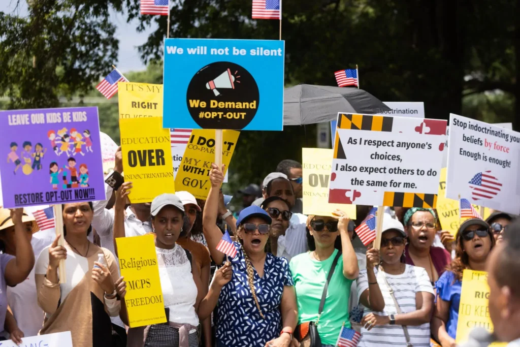 A group of people protest outdoors, holding signs with messages like “We Demand #OPT-OUT,” “Restore OPT-OUT NOW,” and “Stop indoctrinating kids,” raising concerns about Educational Intimidation Bills. Some carry small American flags.