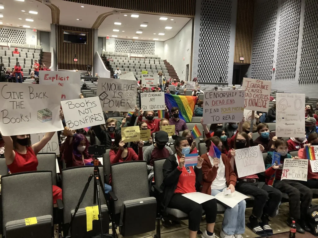 A large group of people sit in an auditorium holding protest signs supporting books and opposing Educational Intimidation Bills, with messages like “Give Back The Books!” and “Knowledge is Power!” Several hold Pride flags.