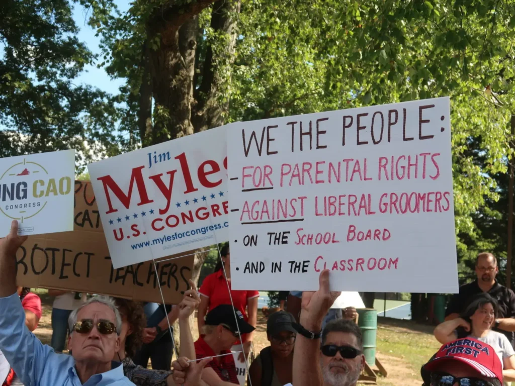 A group of people at an outdoor rally hold up signs, including ones that read “Jim Myle U.S. Congress,” “WE THE PEOPLE: FOR PARENTAL RIGHTS AGAINST LIBERAL GROOMERS,” and others protesting Educational Intimidation Bills.