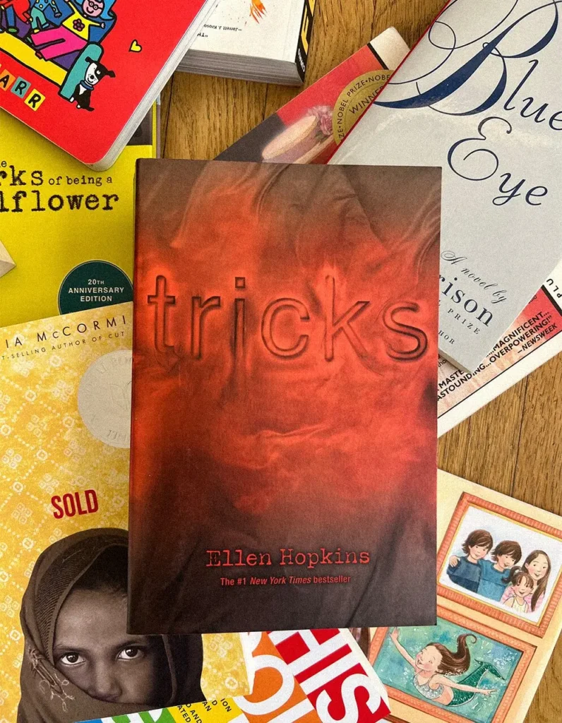 A copy of Tricks by Ellen Hopkins, banned in the USA 2023, sits on a wooden table surrounded by several other books, including Blue Eye, Sold, Works of a Wallflower, and Finding Perfect.