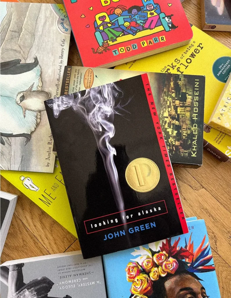 A copy of Looking for Alaska by John Green, banned in the USA 2023, lies on top of a scattered pile of colorful books, including titles by Khaled Hosseini and Todd Parr, on a wooden surface.